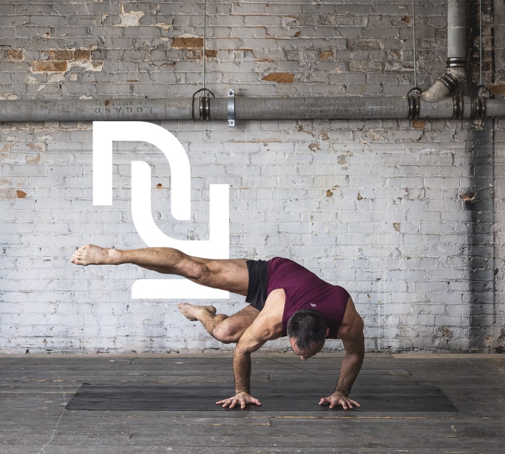 Un homme exécute une posture avancée de power yoga sur les mains, dans un studio au mur de briques.